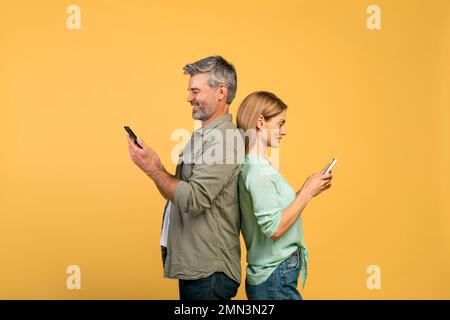 Homme et femme caucasien d'âge moyen debout dos à dos, en utilisant des smartphones, arrière-plan de studio jaune, vue latérale Banque D'Images