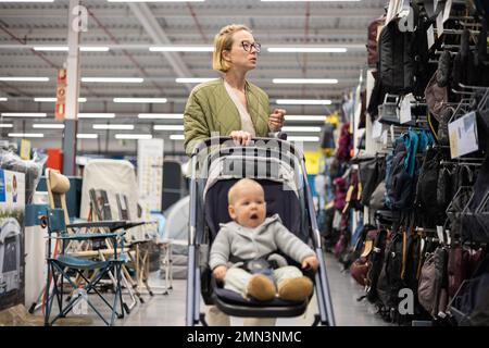 Casualy mère habillée choisissant des chaussures et des vêtements sportifs dans le rayon des sports du magasin de supermarché avec son bébé garçon enfant dans poussette Banque D'Images