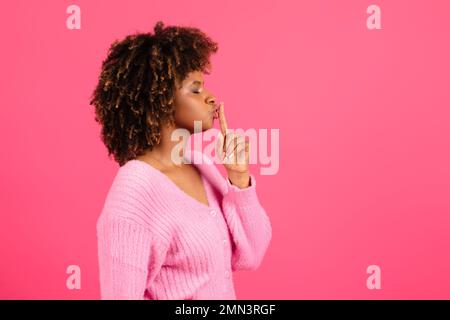 Sérieuse jeune femme noire bouclés dans une ambiance décontractée appuie son doigt sur les lèvres et fait shhh signe Banque D'Images