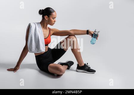 vue latérale de la jeune femme afro-américaine dans un soutien-gorge de sport et un short de vélo assis avec une bouteille de sport et une serviette sur fond gris Banque D'Images