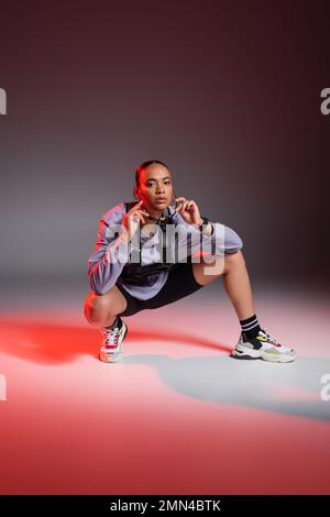 pleine longueur de femme afro-américaine en short de vélo et sweatshirt assis tout en portant des lunettes de soleil sur fond gris avec lumière rouge Banque D'Images