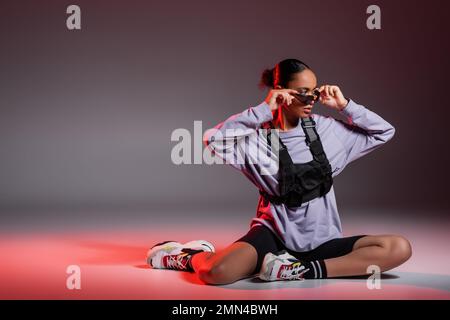 pleine longueur de femme afro-américaine en short de vélo et sweatshirt portant des lunettes de soleil sur fond gris avec lumière rouge Banque D'Images