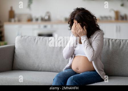 Portrait de la jeune femme enceinte qui pleure en étant assise sur le canapé à la maison Banque D'Images