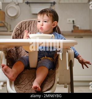 Un enfant drôle mange une pomme râpée avec sa bouche pleine tout en étant assis sur une chaise de cuisine. Bébé garçon affamé, il a de la nourriture dans sa bouche, de l'humour. Enfant âgé Banque D'Images