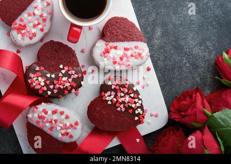 Des biscuits au velours rouge ou au brownie sur le cœur en forme de glaçage au chocolat avec des roses rouges sur fond noir. Idée de dessert pour la Saint-Valentin, les mères ou les femmes Banque D'Images