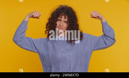 Je suis fort et indépendant. Femme aux cheveux bouclés montrant des biceps, en pleine confiance, sentant la force de pouvoir pour se battre pour les droits, l'énergie pour gagner la réussite gagner. Jeune fille sur fond jaune studio Banque D'Images