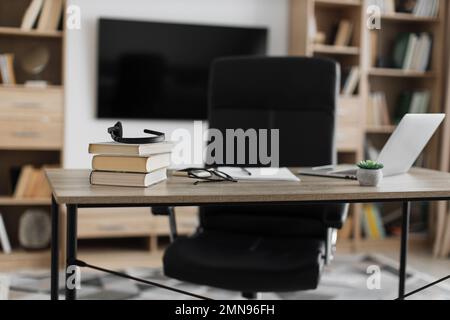 Photo d'un lieu de travail moderne, bureau avec ordinateur portable, livres, papiers, lunettes, micro-casque sur table en bois avec grand téléviseur sur fond de mur blanc. Banque D'Images