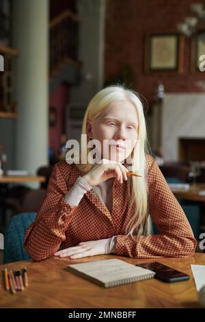 Jeune fille albino dans une tenue décontractée et élégante assis à table dans un café à la lessire et pensant à un nouveau croquis ou s'inspirant de quelque chose Banque D'Images