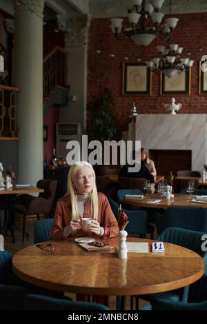 Jeune femme sereine dans de longs cheveux blancs ayant une tasse de thé ou de café tout en étant assis près d'une table dans un café confortable et regardant par la fenêtre Banque D'Images