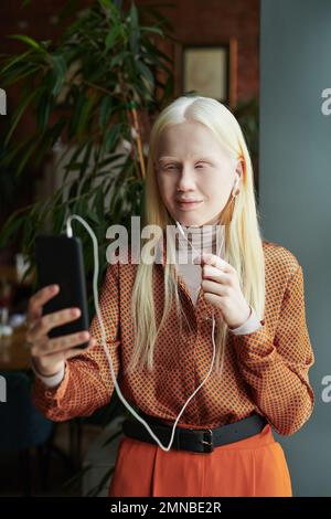 Jeune femme albino dans un chemisier élégant parlant à un ami dans le chat vidéo tout en se tenant devant la caméra contre l'usine verte dans le café Banque D'Images