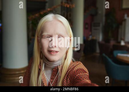 Portrait d'une jeune femme albino avec des écouteurs regardant l'appareil photo tout en tenant le téléphone portable devant son visage et en parlant dans le chat vidéo Banque D'Images