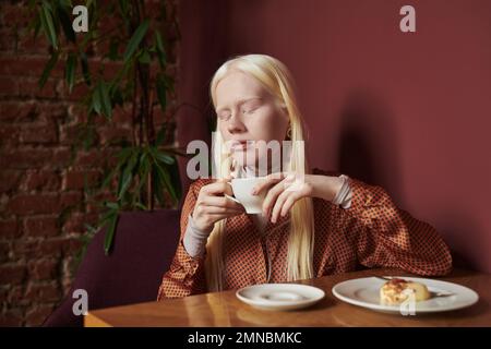 Jeune femme albino sereine avec de longs cheveux blancs tenant une tasse de café tout en étant assis à table avec dessert dans le café avec des plantes vertes Banque D'Images