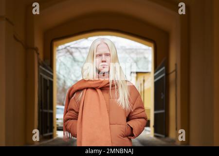 Jeune femme albino élégante avec de longs cheveux très équitables portant une veste et une écharpe marron debout dans un environnement urbain et regardant l'appareil photo Banque D'Images