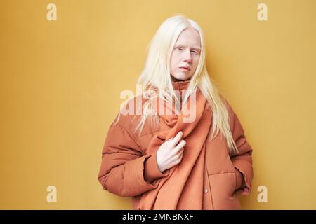 Élégante albino fille dans une veste et une écharpe marron chaud debout sur fond jaune et regardant l'appareil photo tout en posant en isolement Banque D'Images