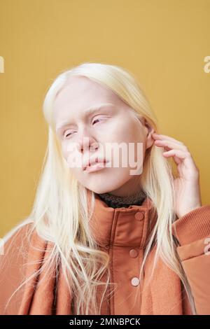 Jeune femme albino sereine avec de longs cheveux blonds debout devant la caméra en isolement contre le mur jaune du bâtiment moderne Banque D'Images