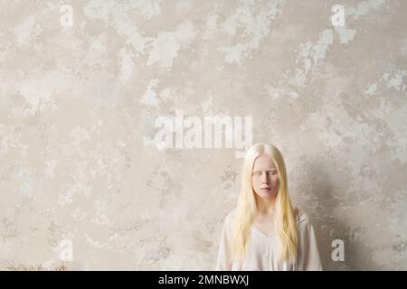 Jeune femme albino sereine en chandail blanc gardant les yeux fermés tout en se tenant par le mur de marbre équitable avec copyspace sur la gauche Banque D'Images