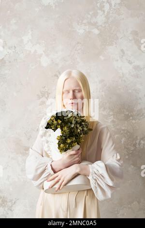 Jeune femme albino sereine en tenue blanche tenant le buste en plastique avec des plantes vertes à l'intérieur tout en se tenant contre le mur avec des papiers peints élégants Banque D'Images