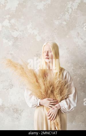 Jeune albino femme avec bouquet de fleurs séchées molletonnées dans les mains en gardant ses yeux fermés tout en se tenant par le mur devant l'appareil photo Banque D'Images