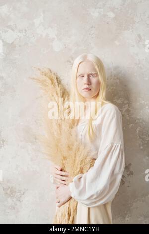 Jeune femme albino sereine en blouse blanche tenant un bouquet de fleurs séchées et regardant la caméra tout en se tenant par un mur blanc Banque D'Images