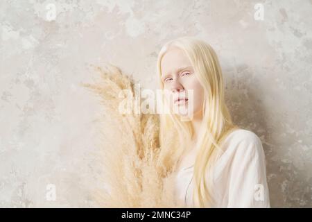 Portrait de la beauté naturelle de paisible albino fille tenant le bouquet de fleurs séchées tout en se tenant près du mur blanc en face de l'appareil photo Banque D'Images