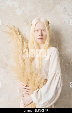 Sereine albino fille avec de longs cheveux tenant un bouquet de fleurs séchées tout en se tenant devant l'appareil photo contre le mur blanc en isolement Banque D'Images