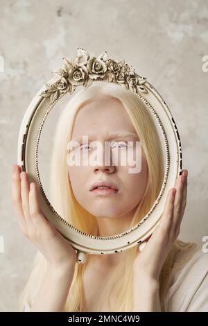 Visage d'une jeune femme albinos sereine dans un cadre ovale antique décoré de roses lors d'une séance photo élégante dans un studio avec des murs en marbre Banque D'Images