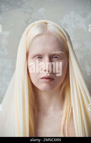 Jeune femme albino sereine avec couverture blanche sur la tête debout devant l'appareil photo contre le mur de marbre pendant la séance photo en studio Banque D'Images