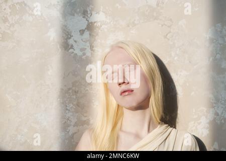 Jeune femme albino sereine en tenue blanche gardant les yeux fermés tout en se tenant près du mur de marbre dans l'isolement et en profitant de la lumière du soleil Banque D'Images