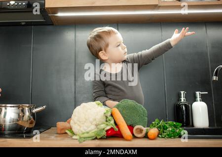 Petit enfant aidant à cuisiner le dîner. Bébé fils assis avec le bras relevé sur le plan d'examen, au milieu d'une pile de légumes frais mûrs. Banque D'Images