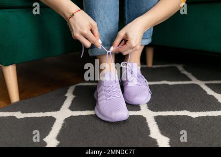 Femme méconnaissable assise sur un canapé vert à la maison, laçage des baskets violettes, étirement des lacets, préparation à la marche. Banque D'Images