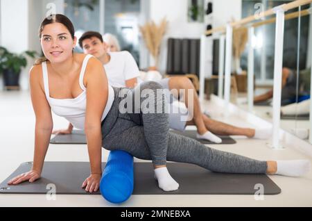 Jeune femme positive effectuant un ensemble d'exercices avec pilates rouleau pendant la classe de groupe dans le studio de fitness. Banque D'Images