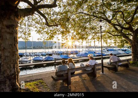 Personnes assises sur des bancs sur la promenade au bord du lac de Zurich, étoile du soleil, soleil du soir, voiliers dans le port derrière, Zurich, Suisse Banque D'Images