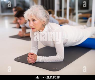 Active senior femme effectue des exercices avec un rouleau pilates tout en étant allongé sur un tapis dans le hall d'un studio de fitness moderne. Banque D'Images