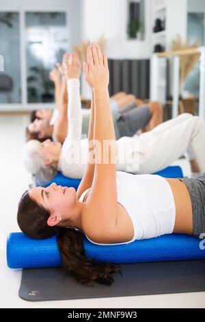 Groupe de personnes engagées dans la forme physique dans la salle de gym exécute un exercice pour améliorer la mobilité du diaphragme, avec rouleau de tapis de sport tordus sous le dos Banque D'Images