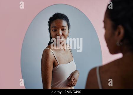 Portrait candid de jeune femme noire avec des cicatrices d'acné regardant dans le miroir et souriant Banque D'Images