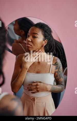 Portrait vertical de la jeune femme noire avec des cicatrices d'acné regardant dans le miroir non sécurisé dans l'apparence Banque D'Images