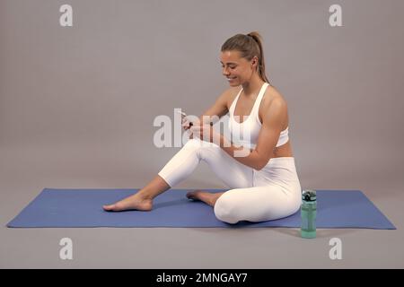 Happy sportswoman à l'aide d'un téléphone portable. Femme de yoga prend des cours de yoga. Une jeune fille sportive paisible dans des vêtements de sport qui s'exercent sur un tapis. Gris isolé Banque D'Images