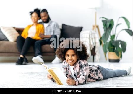 Positive mignon Afro-américaine petite fille avec des cheveux bouclés, se trouve sur le sol dans la salle de séjour, tient un livre dans ses mains, regarde l'appareil photo, sourit, les parents sont assis sur le canapé dans le fond Banque D'Images