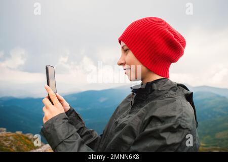 Une jeune femme décontractée hacker dans une casquette rouge prenant des photos dans un smartphone moderne iPhone sur fond d'une chaîne de montagnes pendant un orage Banque D'Images