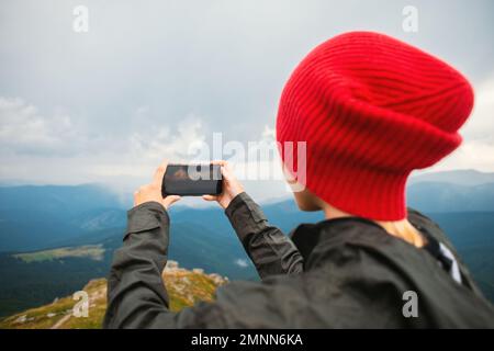 Une jeune femme décontractée hacker dans une casquette rouge prenant des photos dans un smartphone moderne iPhone sur fond d'une chaîne de montagnes pendant un orage Banque D'Images