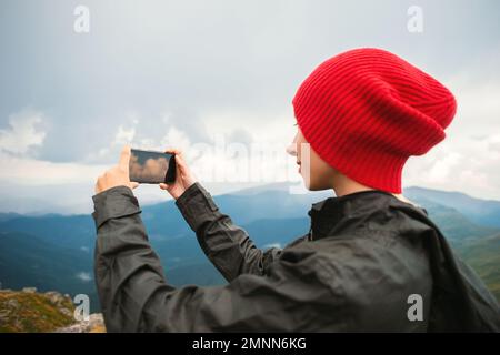 Une jeune femme décontractée hacker dans une casquette rouge prenant des photos dans un smartphone moderne iPhone sur fond d'une chaîne de montagnes pendant un orage Banque D'Images