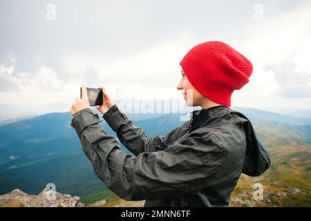 Une jeune femme décontractée hacker dans une casquette rouge prenant des photos dans un smartphone moderne iPhone sur fond d'une chaîne de montagnes pendant un orage Banque D'Images