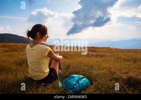 Vue arrière d'une jeune randonneur mignonne se détendre sur le voyage en montagne en vacances sur la montagne en haut regarde son sac à dos contre un ciel nuageux Banque D'Images