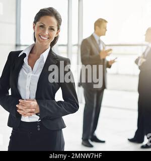 Shes le professionnel de la qualité. Portrait court d'une femme d'affaires debout dans un hall avec ses collègues en arrière-plan. Banque D'Images