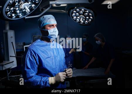 Portrait lumineux de la chirurgie masculine portant un EPI avec des ciseaux chirurgicaux dans les mains debout dans la salle d'opération avec l'équipe médicale Banque D'Images