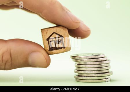 Un homme tient dans sa main un cube en bois représentant une maison près d'une pile de pièces, un concept sur le coût de l'immobilier ou le paiement des taxes Banque D'Images