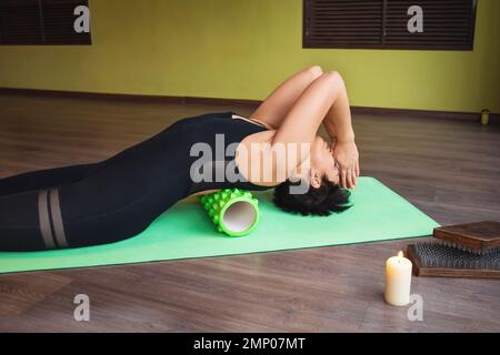 Femme d'âge moyen pratiquant le yoga effectue des exercices relaxants pour les muscles du dos avec un rouleau de massage, s'entraîne avec les yeux fermés tout en étant allongé sur un tapis Banque D'Images