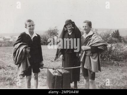 moment vintage / moment drôle vintage / photo vintage / puissance du moment / moments magiques / deux jeunes garçons portent de lourdes valises pour eux grand-mère à la 1930s. Madame porte des vêtements noirs à l'heure d'été. Banque D'Images