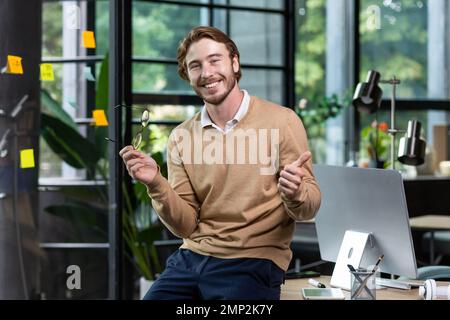Portrait d'un homme blond heureux à l'intérieur du bureau, d'un homme en vêtements décontractés souriant et regardant l'appareil photo, d'un jeune homme d'affaires réussi montrant les pouces à l'appareil photo, d'un programmeur au travail avec un ordinateur. Banque D'Images