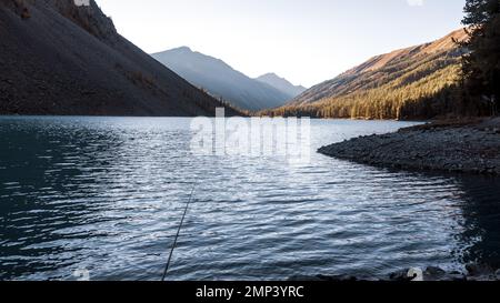 Une canne à pêche se trouve à l'ombre sur les pierres sur la rive du lac alpin Shavlinskoye avec la lumière du soleil le soir dans les montagnes de l'horizontal Banque D'Images